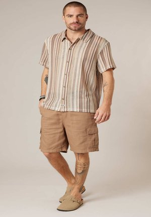 Man met tatoeages, gekleed in een beige gestreept shirt met korte mouwen, bruine cargoshorts en beige instapschoenen, staand met handen in de zakken.