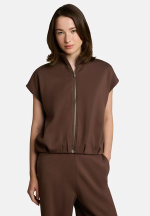 Jeune femme aux cheveux bruns mi-longs, portant un haut marron sans manches à fermeture éclair et un pantalon assorti, debout avec une main dans la poche.