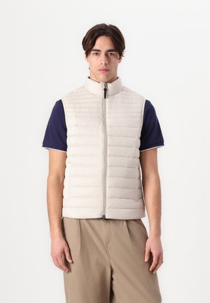Leichte, beige Puffweste mit gesteppter Optik, hohem Kragen und frontalem Reißverschluss. Über einem marineblauen Shirt getragen, kombiniert mit beige Shorts.