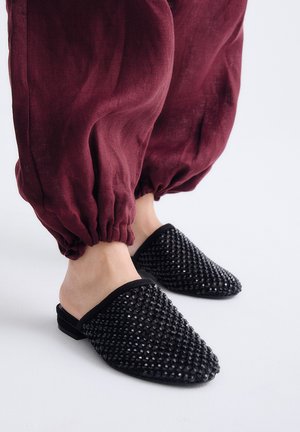 Pies con zapatos tipo mocasín negros con textura, combinados con pantalones sueltos color granate recogidos en los tobillos sobre un fondo blanco.