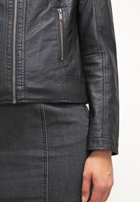 Detailní záběr na osobu, která má na sobě černou koženou bundu s kapsou na zip a tmavě šedou džínsovou sukni, s uvolněnou rukou dosahující k boku.