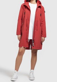 Red waterproof parka with adjustable hood, zipper and toggle closures, two side pockets, and a relaxed fit, worn over a white shirt and shorts.