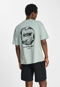 Ljusgrön t-shirt med kort ärm som har en svart fotbollsgraphic och texten "HSW" och "serve the game" på baksidan. Tillverkad av bomull.