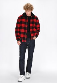 Rot-schwarz karierte Flanelljacke mit schwarzem Kragen, Reißverschluss, kombiniert mit dunklen Jeans und weißen Sneakers. Darunter ein schlichtes schwarzes T-Shirt.