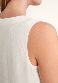 Weißes ärmelloses Leinenoberteil mit abgerundetem Ausschnitt, einer strukturierten Oberfläche und einem lockeren Sitz, das Schulter- und oberen Rückenbereich zeigt.