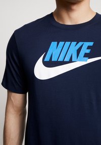 Marinblå t-shirt med en stor vit Nike-swoosh och stiliserad blå "NIKE" text. Kurvade ärmar, rund halsringning, mjukt tyg.