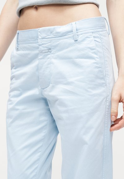 Κοντινό πλάνο ενός ατόμου που φοράει ανοιχτό μπλε παντελόνι και cropped γκρι τοπ, εμφανίζοντας τη μέση, τους γοφούς και το δεξί χέρι, με φόντο καθαρό λευκό.