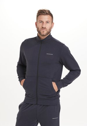 Marineblå zip-up sportsjakke med ståkrave, to forlommer og "ENDURANCE" logo på brystet. Glat stof med en tætsiddende pasform.