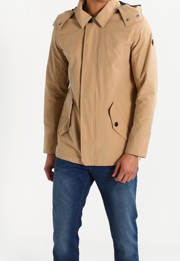 Homme portant une veste beige à capuche avec des poches à rabat boutonnées et un jean bleu, debout devant un fond blanc uni.