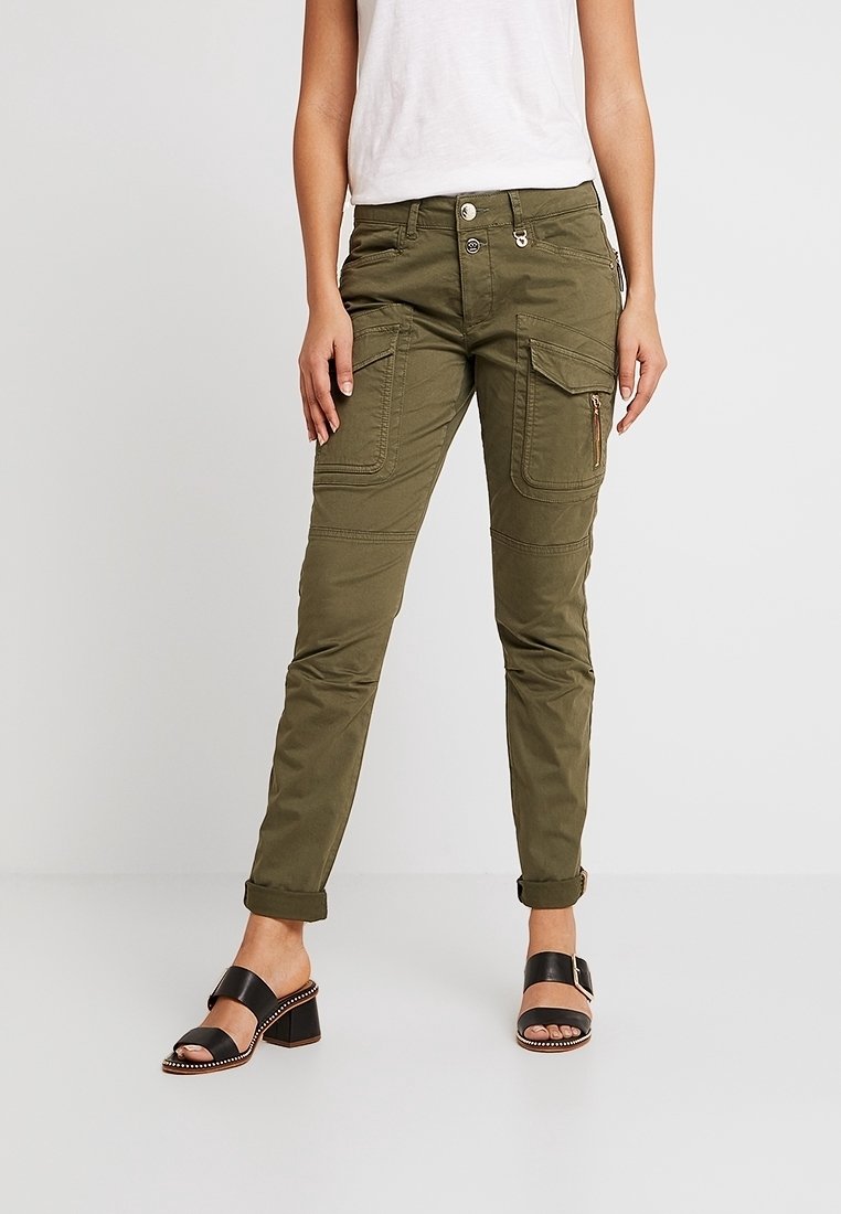 Personne portant un pantalon cargo vert olive avec de grandes poches avant et des sandales à talons blocs noires à bout ouvert, debout devant un fond uni.