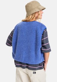 Blå fleecevest med en teksturert overflate, med blå kant. Båret over en mørk stripete skjorte. Tilbehør: beige bøttehatt.