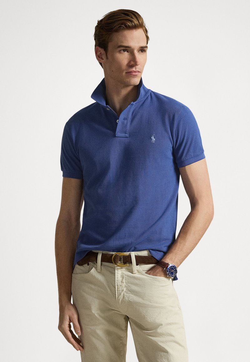 Blue polo shirt with a folded collar, two buttons, and a small logo. Paired with beige trousers and a brown belt. Features a watch on wrist.
