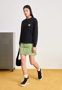 Schwarzes Langarmhemd mit einer Brusttasche, kombiniert mit einem grünen Rock, schwarzen High-Top-Sneakern und einer schwarzen Tasche. Neutraler Hintergrund.