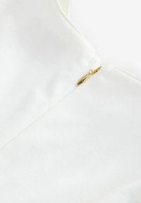 Top blanco con textura suave, diseñado con un escote en V poco profundo y cierre de cremallera dorada en el lado izquierdo.