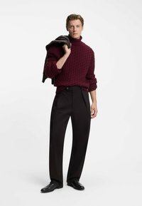 Bordeaux kabelgebreide trui met hoge hals, zwarte wijde broek en zwarte leren schoenen. Model houdt een bruine jas over de schouder.