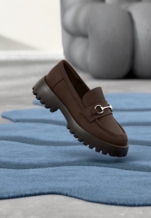 Mocassin marron foncé avec semelle épaisse rainurée et détail argenté reposant sur un tapis bleu texturé avec un mobilier gris rembourré en arrière-plan.