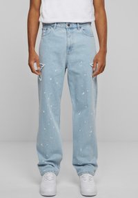 Light blue distressed denim jeans featuring paint splatter details, straight leg design, and frayed knee accents, paired with white sneakers.
