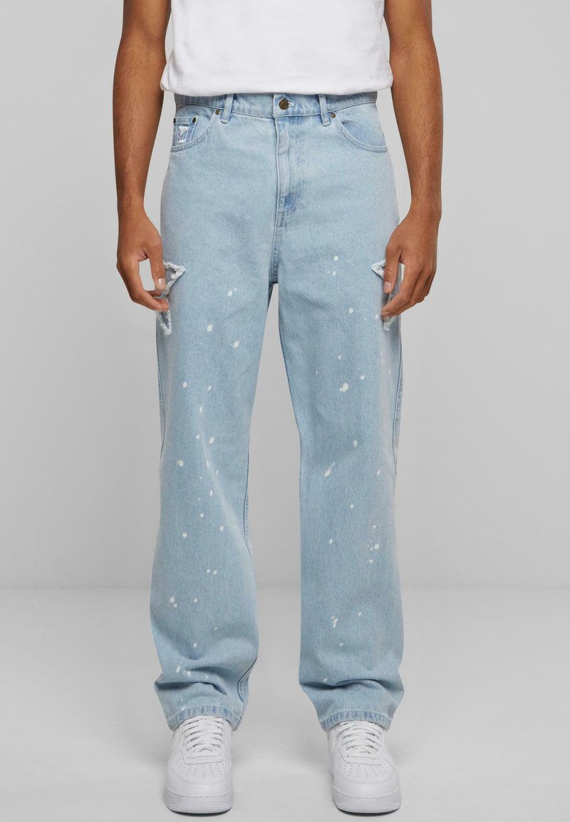 Light blue distressed denim jeans featuring paint splatter details, straight leg design, and frayed knee accents, paired with white sneakers.