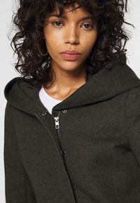 Veste à capuche verte en tissu texturé, dotée d'une fermeture éclair et de boutons-pression. Portée sur une chemise blanche, avec un ajustement serré autour du cou.