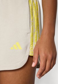Saia atlética de tom bege claro com acabamento amarelo e padrão subtil. Apresenta o logótipo da Adidas em amarelo. Textura suave com um ligeiro brilho.