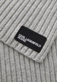 Pilkos megztos medžiagos su vertikaliomis raukšlėmis ir tekstūruota juoda etikete, ant kurios balta siuvinėjimu užrašyta "KARL LAGERFELD JEANS".