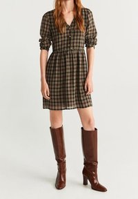 Braun kariertes Kleid mit tailliertem Schnitt, Knopfleiste und Puffärmeln, kombiniert mit hohen braunen Lederstiefeln.