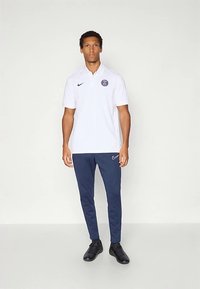 Muž stojí oblečený v bielom polo tričku PSG, námorníckych nike teplákoch a čiernych teniskách na jednolítom svetlom pozadí.