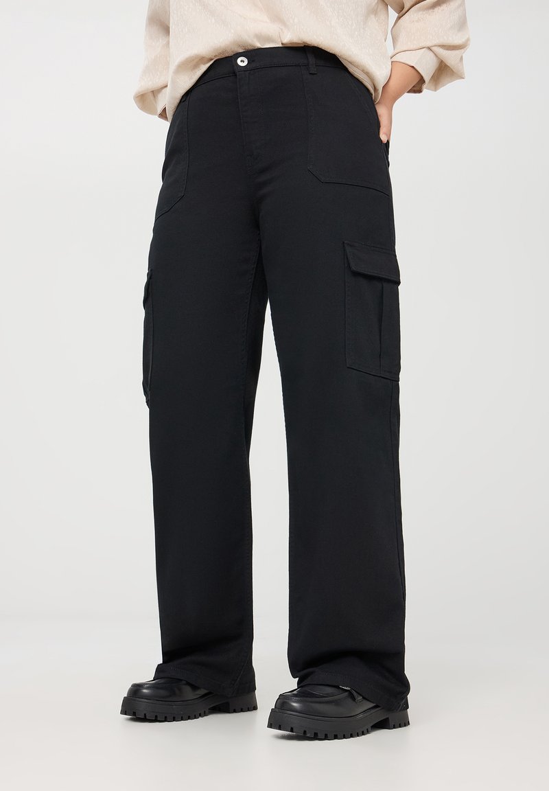 Pantaloni cargo neri realizzati in tessuto spesso, dotati di tasche laterali, design a gamba dritta e cuciture rinforzate. Indossati con scarpe nere chunky.