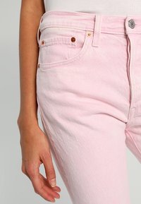 Vaqueros de mezclilla rosa claro con un diseño clásico. Las características incluyen cinco bolsillos, herrajes de color cobre y una costura del cinturón visible.