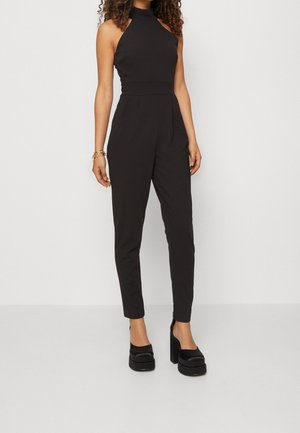 Czarny kombinezon o wysokim dekolcie i bezrękawowym kroju, dopasowanej górze, zwężanych nogawkach i teksturowanej tkaninie. W połączeniu z masywnymi czarnymi butami na platformie.
