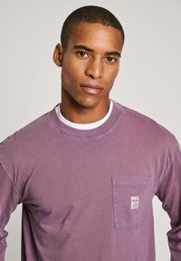 Long-sleeved, crew neck shirt in light purple cotton, featuring a chest pocket with a small logo label and visible stitching details.