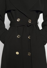 Manteau noir croisé avec des boutons dorés, une ceinture en tissu nouée à la taille, et les mains glissées dans les poches latérales.