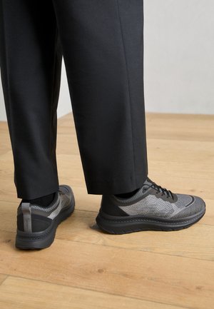 Scarpe da ginnastica grigie con una tomaia in mesh testurizzato, dettagli neri e una suola spessa, abbinate a pantaloni neri su misura su un pavimento di legno.