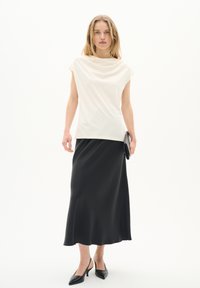 Haut drapé crème avec des manches courtes, associé à une jupe midi en satin noir. Le mannequin tient une pochette noire et porte des escarpins noirs pointus.
