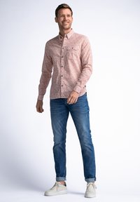 Camisa de botones de color rosa claro con estampado floral y un bolsillo en el pecho, combinada con jeans azules y zapatillas blancas. Textura suave, diseño casual.