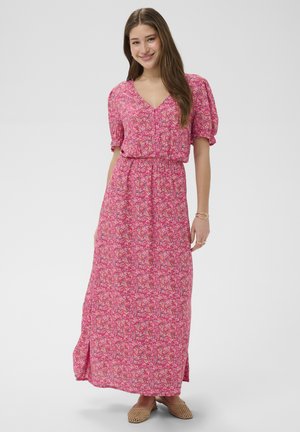 Jonge vrouw die glimlacht, gekleed in een roze maxi-jurk met bloemenprint, V-hals en pofmouwen, met een elastische taille en beige gevlochten platte schoenen.