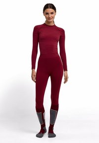 Vrouw staand in bijpassende naadloze, bordeauxrode lange mouwen top en legging met grijze gewatteerde sokken, poserend tegen een witte achtergrond.