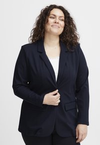 Jūros mėlynumo švarkas su glotnia tekstūra, vienu mygtuku, įpjovomis apykaklėmis ir dviem priekinėmis kišenėmis. Užsidėtas ant balto marškinėlių su nėrinių apdaila.