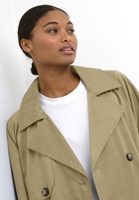 Beige Trenchcoat mit großem Kragen, doppelt geknöpftem Verschluss und glatter Textur, über einem weißen T-Shirt getragen.