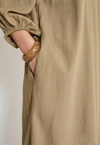 Khakikleid mit gepufften Ärmelbündchen, versehen mit einer seitlichen Tasche. Ein glänzendes, tanfarbenes Armband schmückt das Handgelenk und hebt den strukturierten Stoff hervor.