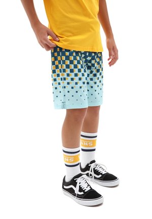 Garçon portant un short à carreaux bleu et jaune, un t-shirt jaune, des chaussettes blanches Vans et des baskets noires Vans, debout devant un fond blanc.