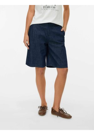 Person iført mørkeblå knælange denimshorts, hvid ærmeløs top og brune snørestøvler, stående med hænderne nær lommerne.