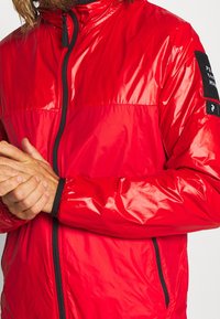 Rød letvægtsjakke lavet af skinnende materiale; har en fuld lynlås foran og sorte detaljer på ærmerne. Tekstureret, puffet design.