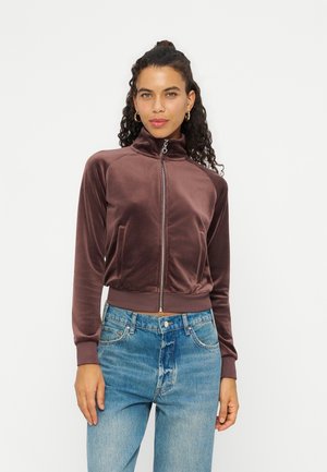Femme aux cheveux bouclés portant une veste en velours marron zippée et un jean bleu, se tenant devant un fond blanc uni.