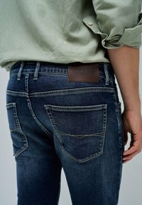 Tamno plave traperice s uskim krojem, s kožnom oznakom na kojoj piše "SALSA JEANS" i karakterističnim šavovima na stražnjim džepovima.