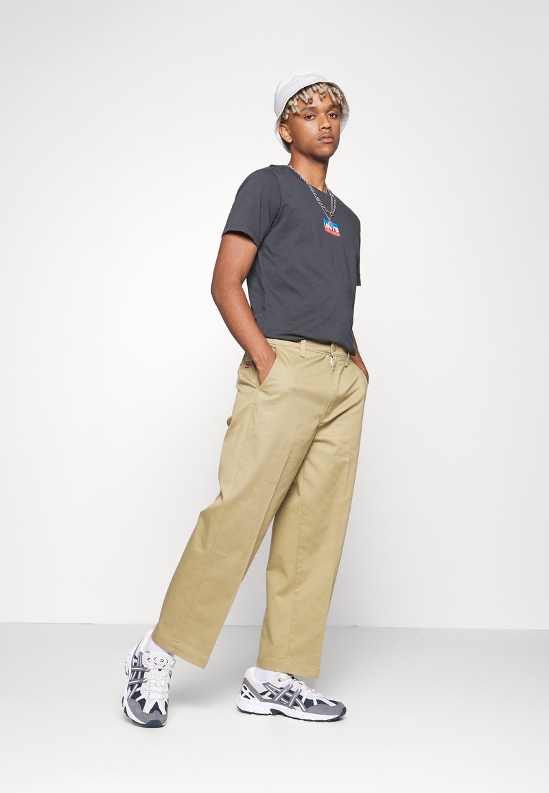 Outfit casual composto da una t-shirt grigia con logo, pantaloni khaki e sneaker bianche con dettagli grigi. Il modello indossa un cappello a secchiello bianco.