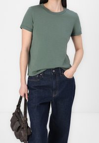 Zielona bawełniana koszulka z krótkimi podwiniętymi rękawami, zestawiona z ciemnoniebieskimi spodniami z wysokim stanem i brązową skórzaną torbą.