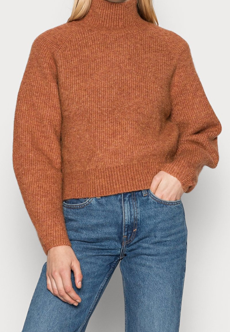 Personne portant un pull en maille côtelée couleur rouille et un jean taille haute bleu, avec une main dans la poche, sur un fond uni.