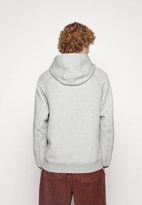 Persona con cabello rizado vistiendo una sudadera gris claro y pantalones de pana marrón, mostrada desde la parte trasera contra un fondo blanco liso.