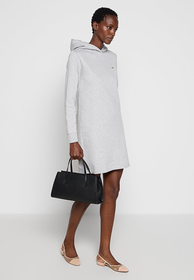 Un vestido de sudadera gris con mangas largas, aberturas laterales y un pequeño logo. Combinado con un bolso negro y unas bailarinas beige para un look casual.